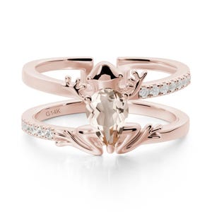 May include: Rose gold ring with a frog design and a pear-shaped gemstone. The ring has a split shank design, with one band set with small, clear stones. The inscription "G14K" is visible on the band.