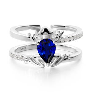 May include: A silver ring with a large, pear-shaped, deep blue gemstone at the centre. The ring features a deer antler design and a band with small, clear stones. The inscription "G14K" is visible on the band.