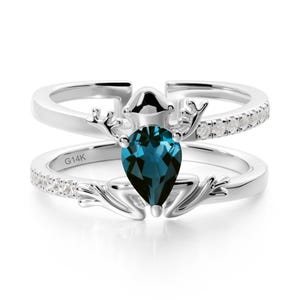 May include: A two-piece silver ring set. The top ring features a row of small, clear stones. The bottom ring has a large, pear-shaped, teal-coloured gemstone. The rings are connected by a decorative element. The bottom ring is marked with "G14K".