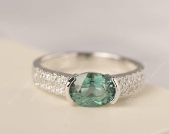 May include: A silver ring featuring a large, oval-cut, teal-green gemstone. The band is adorned with small, clear accent stones on either side of the main stone, creating a sparkling effect. The ring is set against a neutral background.