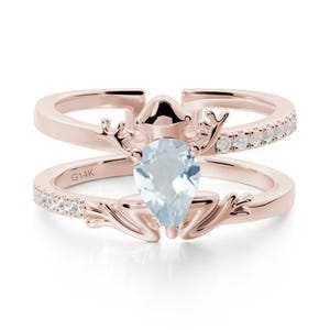 May include: Rose gold ring featuring a pear-shaped, light blue gemstone. The ring has a distinctive frog design, with small white gemstones set along the band. The inscription "G14K" is visible on the ring.
