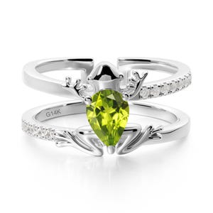 May include: A silver ring featuring a pear-cut, light green gemstone. The ring has a double band design, with small clear stones set along the top band. The inscription "G14K" is visible on the lower band.