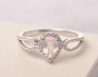 May include: A silver ring featuring a pear-shaped, light pink gemstone surrounded by a halo of small, clear stones. The band has a split design with additional small stones, creating a sparkling effect.