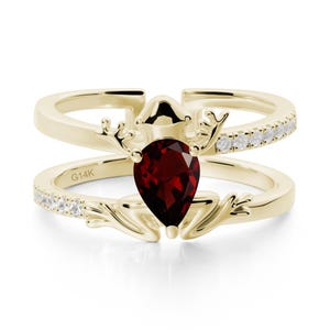 May include: A two-piece gold ring set. The top ring features a row of small, clear stones. The bottom ring has a pear-shaped, deep red gemstone. The rings are connected by a decorative element. The bottom ring is stamped with "G14K".