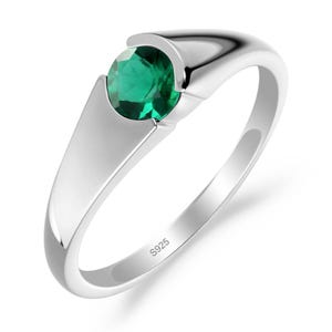 Peut inclure: Une bague en argent poli avec une pierre précieuse verte ronde et facettée, montée dans un style tension. L'anneau est lisse et effilé, avec le poinçon S925 clairement gravé sur la surface intérieure du métal, indiquant de l'argent sterling.