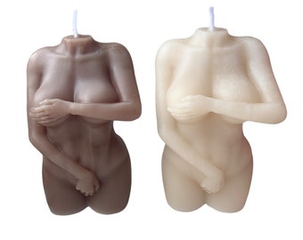 May include: Two nude-colored candles shaped like the female form. One is a light brown color, and the other is a creamy off-white. Both candles have a hand covering the breasts and are headless, with a wick extending from the neck.