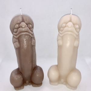 May include: Two novelty candles shaped like a stylized figure with a head and phallic body. One candle is a light brown color, and the other is off-white. Both candles have a wick at the top.