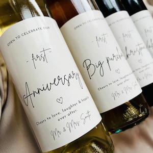 May include: Close-up of four wine bottles with custom labels. The labels read "First Anniversary", "Big Purchase", and "Christmas". Each label includes a heart symbol and the phrase "Cheers to love, laughter & happily ever after."