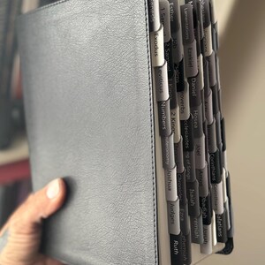 May include: A gray leather Bible with labeled tabs. The tabs are in shades of gray and white, with text indicating the books of the Bible. The cover has a textured appearance. The book is open, revealing the pages and tabs.