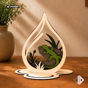 May include: A light-colored wooden cutout in the shape of a teardrop featuring a layered design. Inside the frame, a bright green lizard is positioned on a dark gray rock, surrounded by dark green foliage elements on a flat wooden base.