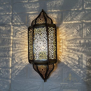 Puede incluir: Una lámpara de pared de estilo marroquí con intrincados trabajos en metal y una luz cálida y brillante. La lámpara está hecha de latón y tiene un diseño tradicional con patrones geométricos. Está montada en una pared de ladrillo blanco.