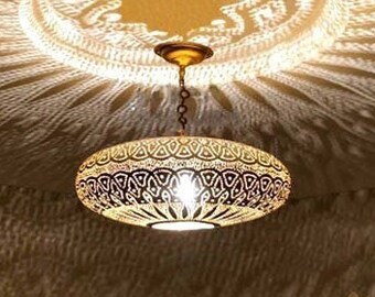 May include: A gold Moroccan pendant light fixture with intricate cutouts casts a decorative shadow on the ceiling. The light fixture is hanging from a chain and has a round, flat shape. The shadow is a repeating pattern of circles and stars.