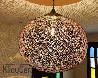 May include: A round, Moroccan-style pendant light fixture with a detailed, swirling pattern. The light casts intricate shadows on the ceiling. The fixture is suspended from a chain and features the text "KliouCera Moroccan Lighting."
