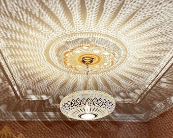 May include: A gold-toned chandelier with a crystal design hangs from a ceiling with a decorative pattern. The chandelier casts a geometric pattern of light on the ceiling.