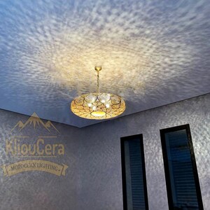May include: A gold Moroccan pendant light fixture with a circular design hangs from a white ceiling. The light fixture casts a pattern of light on the white ceiling. The room has two windows with black frames and white blinds. The walls are a light grey colour.