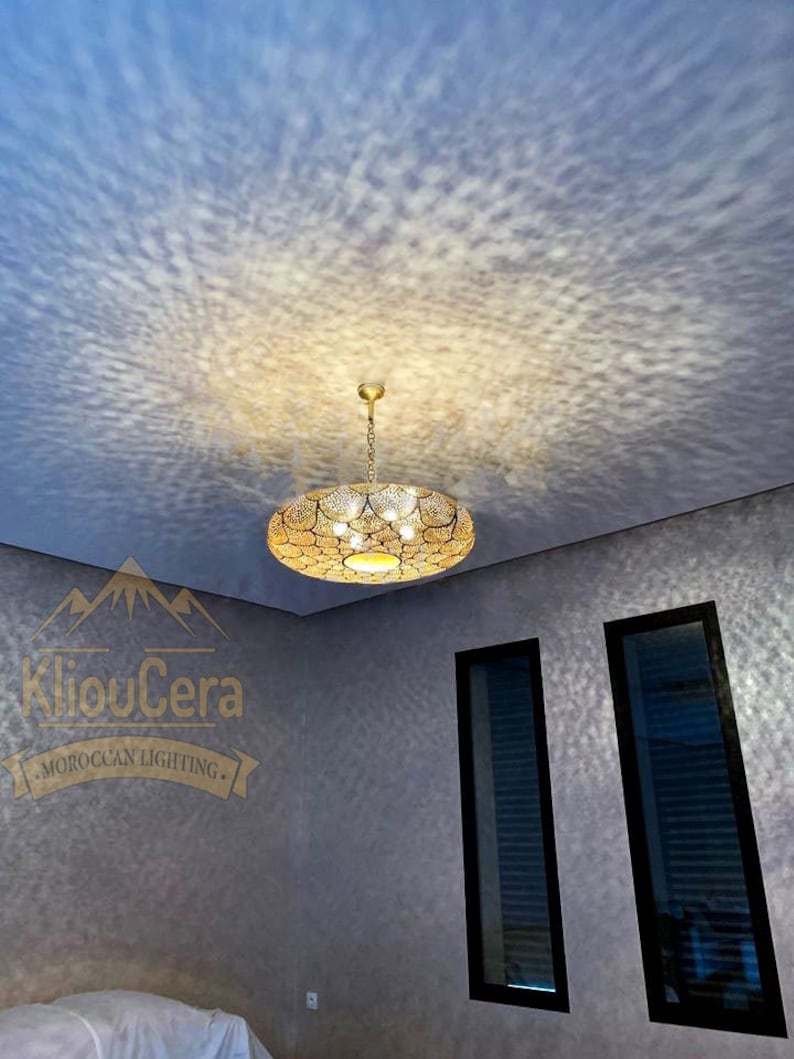May include: A gold Moroccan pendant light fixture with a circular design hangs from a white ceiling. The light fixture casts a pattern of light on the white ceiling. The room has two windows with black frames and white blinds. The walls are a light grey colour.