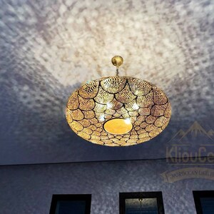 May include: A large, round, gold-coloured Moroccan pendant light fixture with a decorative, geometric pattern. The light fixture is hanging from a ceiling and is casting a shadow on the wall.