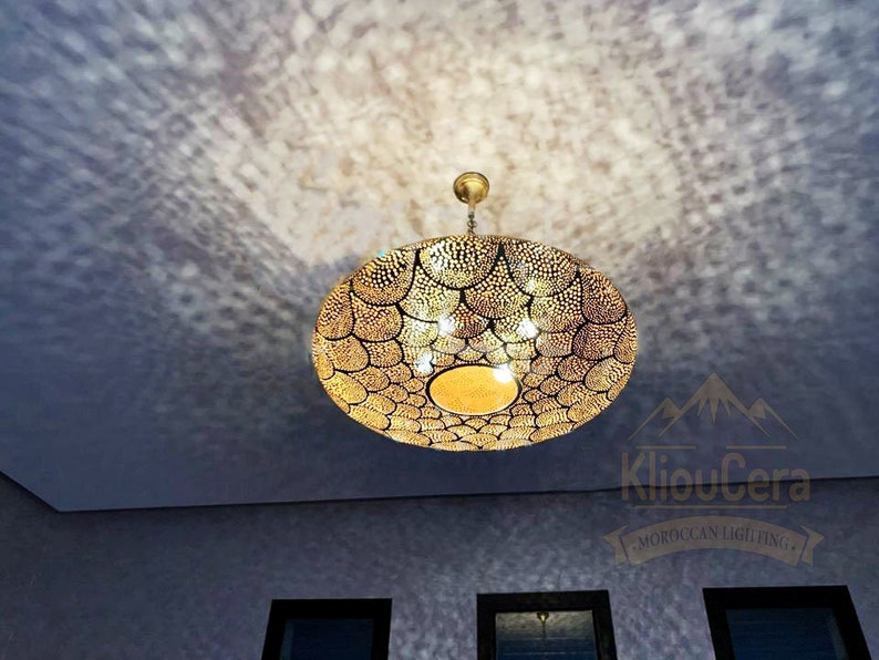 May include: A large, round, gold-coloured Moroccan pendant light fixture with a decorative, geometric pattern. The light fixture is hanging from a ceiling and is casting a shadow on the wall.