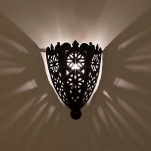Könnte beinhalten: Eine schwarze Wandleuchte aus Metall mit filigranen Ausschnitten. Die Leuchte ist beleuchtet und wirft einen Schatten auf die Wand dahinter.