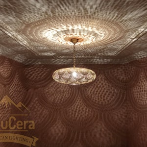 May include: A gold Moroccan pendant light fixture with a round, openwork design hangs from a ceiling. The light casts intricate patterns on the wall, creating a decorative effect.