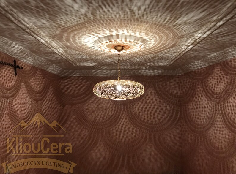 May include: A gold Moroccan pendant light fixture with a round, openwork design hangs from a ceiling. The light casts intricate patterns on the wall, creating a decorative effect.