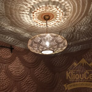 May include: A round, brass Moroccan pendant light with intricate cut-out designs. The light casts a decorative shadow pattern on the ceiling. The light is hanging from a chain and has a simple, round ceiling fixture.