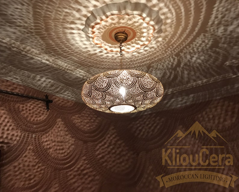 May include: A round, brass Moroccan pendant light with intricate cut-out designs. The light casts a decorative shadow pattern on the ceiling. The light is hanging from a chain and has a simple, round ceiling fixture.