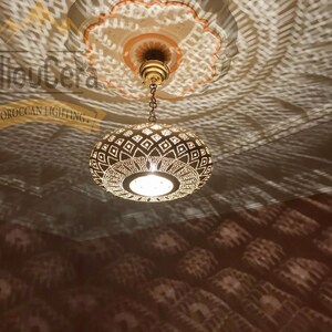 May include: A gold Moroccan pendant light with a geometric pattern hangs from a chain. The light casts intricate shadows on the ceiling.