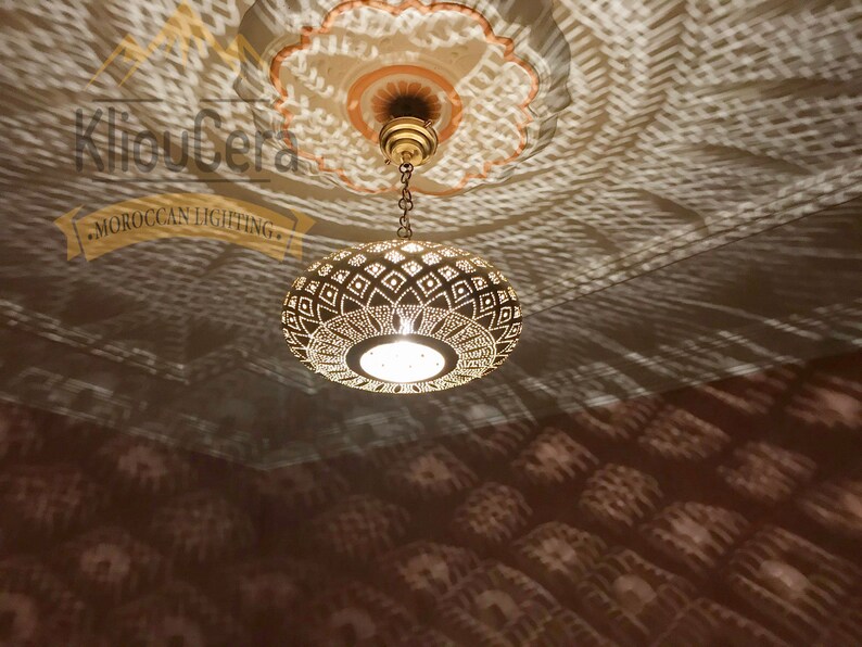 May include: A gold Moroccan pendant light with a geometric pattern hangs from a chain. The light casts intricate shadows on the ceiling.