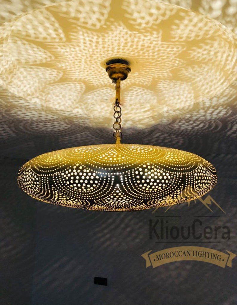 May include: A gold-coloured, oval-shaped pendant light fixture with a decorative, intricate pattern of small holes. The light fixture is hanging from a chain and is casting a patterned shadow on the ceiling.