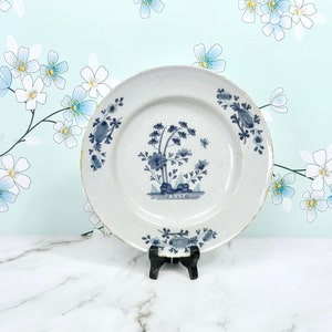 May include: A white ceramic plate with a blue and white floral design. The plate is on a white marble surface and has a black stand.