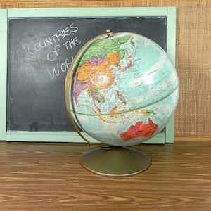 Może przedstawiać: Antyczny globus z niebieską i zieloną mapą świata. Globus stoi na metalowej podstawie i ma mosiężny południk. Globus znajduje się przed tablicą z napisem "Countries of the World".