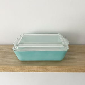 May include: A rectangular turquoise glass baking dish with a clear glass lid. The dish features integrated handles on both sides and a smooth, opaque finish. The lid is transparent with a raised rectangular center section and a flat rim.