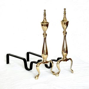 May include: A pair of fireplace andirons featuring polished brass vertical finials with a tapered, urn-shaped design. Each brass finial is supported by three curved legs and attached to a long, black iron horizontal bar extending backward.