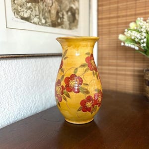 Puede incluir: Un jarrón de cerámica amarillo con un diseño floral. El jarrón tiene flores rojas y verdes pintadas.