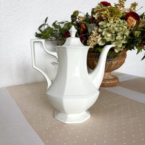 May include: A white porcelain coffee pot with a geometric design. The pot has a tall, slender spout and a handle with a rectangular opening. The lid is topped with a small finial. The pot is displayed on a beige and white patterned surface.