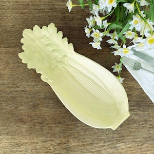 May include: A pale yellow ceramic serving platter shaped like a leaf of lettuce. The platter has detailed veining and a textured surface. White flowers and silverware are in the background.