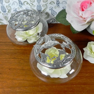 Pode incluir: Dois frascos de vidro transparente com tampas prateadas com desenhos de beija-flores. Cada frasco contém uma rosa branca.