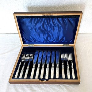 May include: A wooden storage box with a hinged lid, lined with dark blue velvet, containing a set of twelve pieces of flatware. The set includes six silver-toned forks and six silver-toned knives, all featuring white marbled handles with metallic accents.