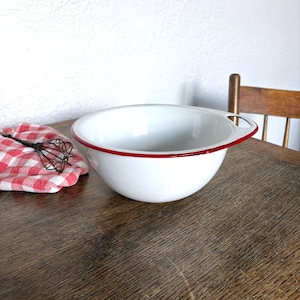 May include: A white enamel mixing bowl with a red rim and a single side handle, accompanied by a black wire whisk resting on a red and white chequered cloth. The bowl features a smooth, glossy finish and a deep, rounded basin for food preparation.