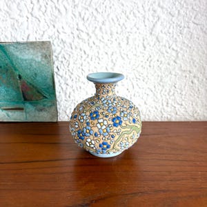 May include: A small, round ceramic vase with a light tan base and a pattern of blue and white flowers. The vase has a light blue rim and a slightly textured surface. A small painting is in the background.