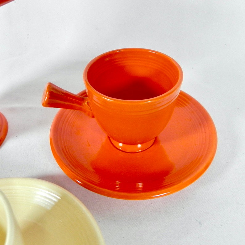 May include: A bright orange ceramic teacup and saucer set. The cup has a unique handle and a rounded shape. The saucer has a simple design with a slight lip.