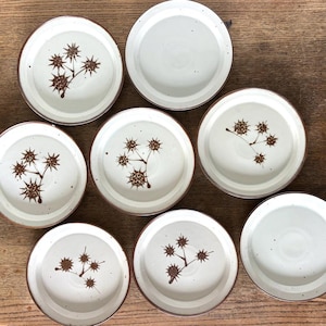 May include: A set of seven round ceramic plates with a cream-colored glaze and brown speckled details. Five of the plates feature a brown hand-painted botanical illustration of spiky seed pods on a stem, while two plates remain plain.