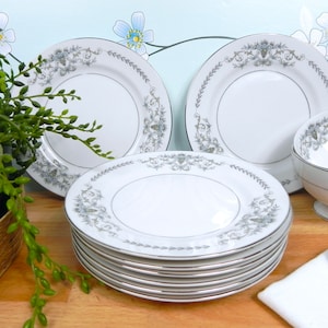 Peut inclure: Un ensemble de dix assiettes plates en céramique blanche avec un bord argenté et un motif floral délicat. Les assiettes sont empilées sur une surface en bois.