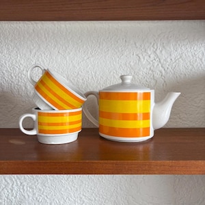 Könnte beinhalten: Ein Set aus weißen Keramik-Teetassen und einer Teekanne mit orangefarbenen und gelben Streifen. Die Tassen sind übereinander gestapelt und die Teekanne steht auf einem Holzregal.