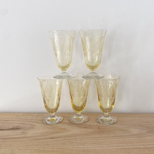 May include: Six vintage yellow-tinted stemmed glasses. Each glass has a delicate floral pattern etched into the glass. The glasses are arranged in a pyramid shape on a wooden surface, with a white background.