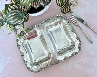 Pode incluir: Uma travessa prateada com dois compartimentos retangulares. A travessa tem uma borda decorativa com um desenho de videira. Um garfo e uma faca são colocados ao lado da travessa. Uma planta em vaso está no fundo.