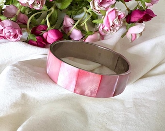 May include: A pink and silver bangle bracelet with a rectangular design. The bracelet is made of pink mother-of-pearl segments and a silver-tone metal band. The bracelet is displayed on a white cloth with pink roses in the background.