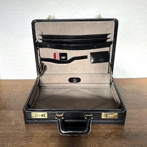May include: Open black leather briefcase with gold-tone hardware. The interior has multiple pockets, a card holder with a red card, and a tan lining. The briefcase has a combination lock and a handle.