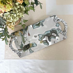 Puede incluir: Una bandeja de servir plateada de forma rectangular con dos asas ornamentadas. La bandeja tiene una textura martillada, que refleja la luz y crea un efecto ondulado. Las asas tienen un diseño de cuerda trenzada. Adecuada para servir alimentos o exhibir objetos.